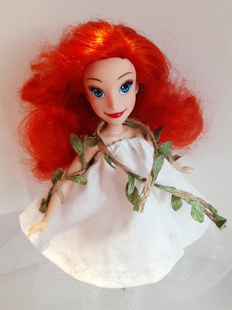Puede incluir: Una mu&ntilde;eca de pelo rojo con un vestido blanco con hojas verdes y cuerda alrededor de la cintura. La mu&ntilde;eca tiene ojos azules y una cara sonriente.