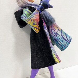 May include: A fashion doll in a black glittery dress with a colorful patterned shawl and purple boots. The doll has lavender hair, purple sunglasses, and blue gloves. The doll is posed with one arm raised.