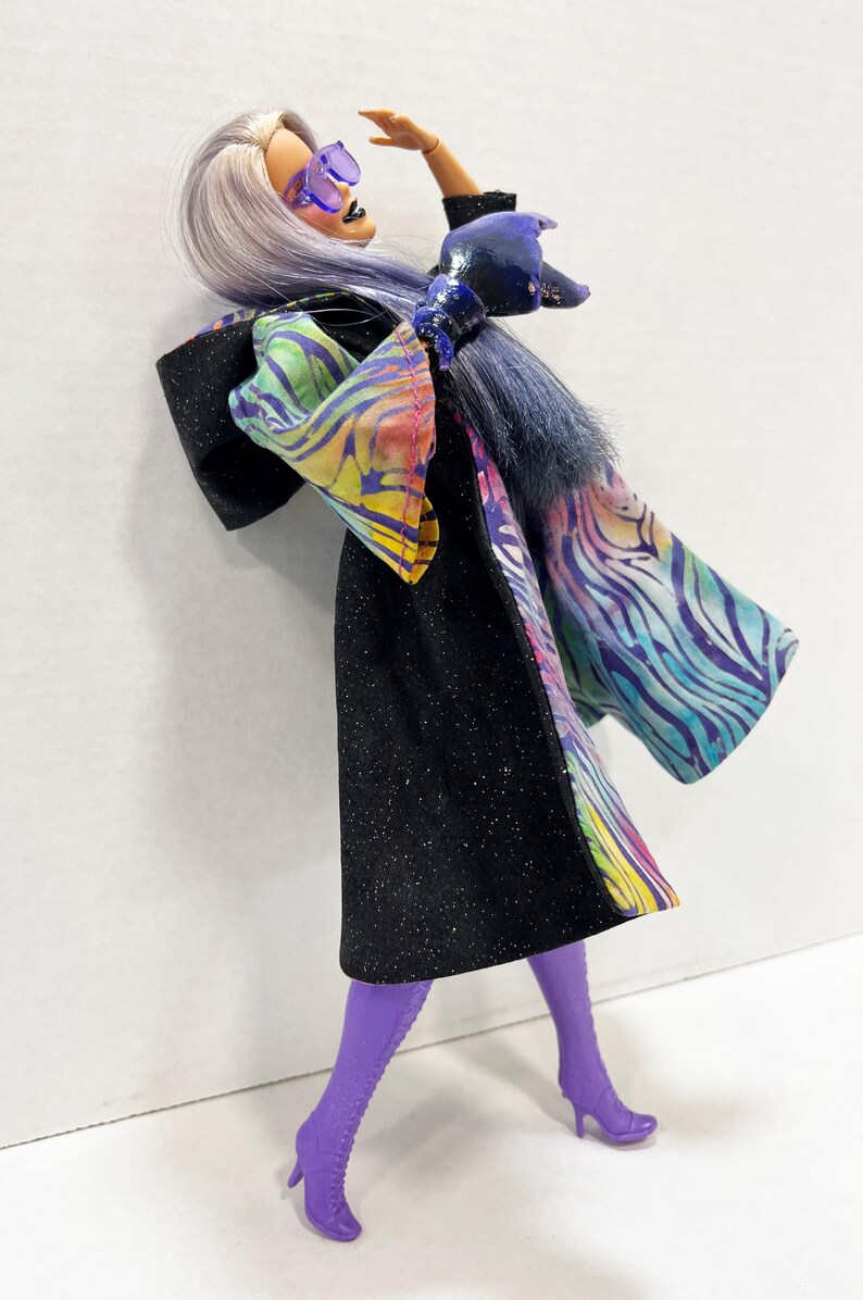 May include: A fashion doll in a black glittery dress with a colorful patterned shawl and purple boots. The doll has lavender hair, purple sunglasses, and blue gloves. The doll is posed with one arm raised.