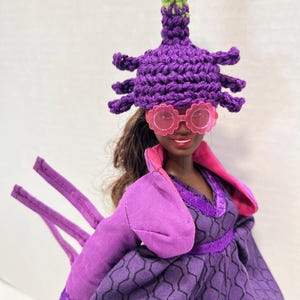 May include: A doll dressed in a purple outfit, including a crocheted hat with a pink and green flower, and pink sunglasses. The dress has a geometric pattern and a pink collar. The doll has brown hair.