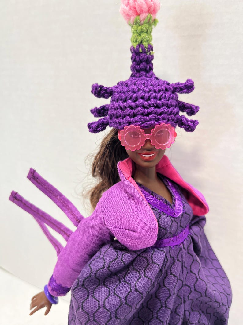 May include: A doll dressed in a purple outfit, including a crocheted hat with a pink and green flower, and pink sunglasses. The dress has a geometric pattern and a pink collar. The doll has brown hair.