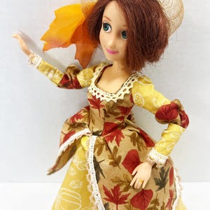 May include: A doll with auburn hair wearing a straw-colored hat and a dress featuring a tan bodice with red and brown autumn leaf patterns, a yellow skirt with white pumpkin outlines, and white lace trim along the neckline and skirt edges.
