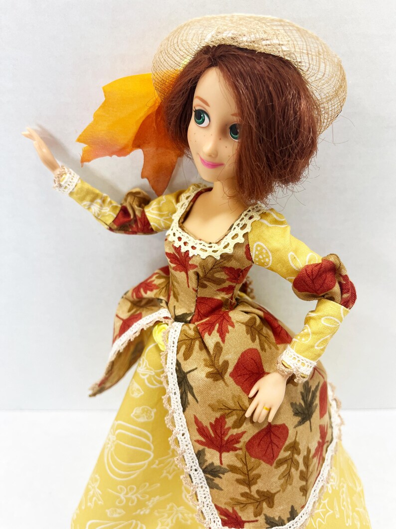 May include: A doll with auburn hair wearing a straw-colored hat and a dress featuring a tan bodice with red and brown autumn leaf patterns, a yellow skirt with white pumpkin outlines, and white lace trim along the neckline and skirt edges.