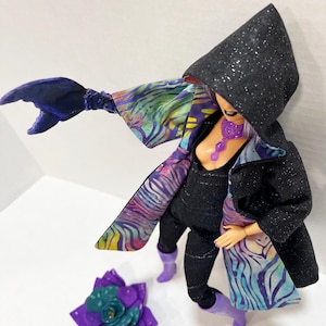 May include: A doll dressed in a black hooded coat with silver glitter, black jumpsuit, and purple boots. The coat has a colorful patterned lining. A purple flower and a claw-like arm are also present.