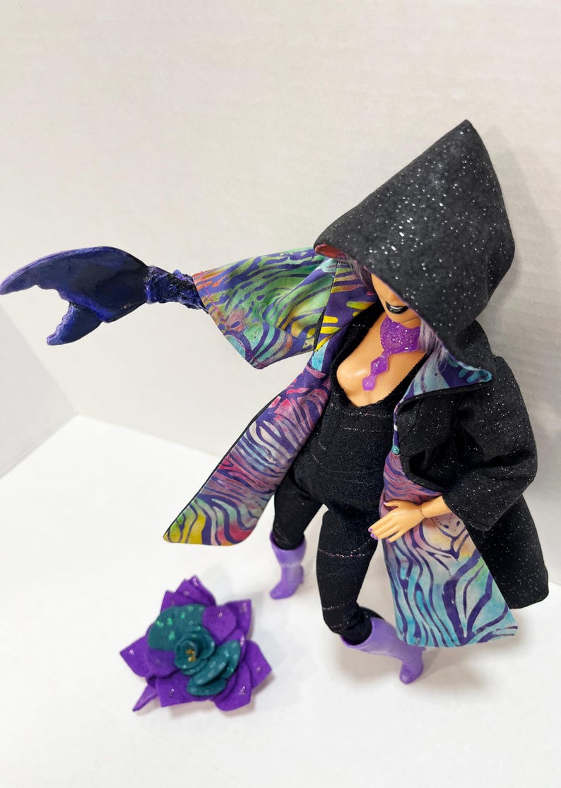May include: A doll dressed in a black hooded coat with silver glitter, black jumpsuit, and purple boots. The coat has a colorful patterned lining. A purple flower and a claw-like arm are also present.