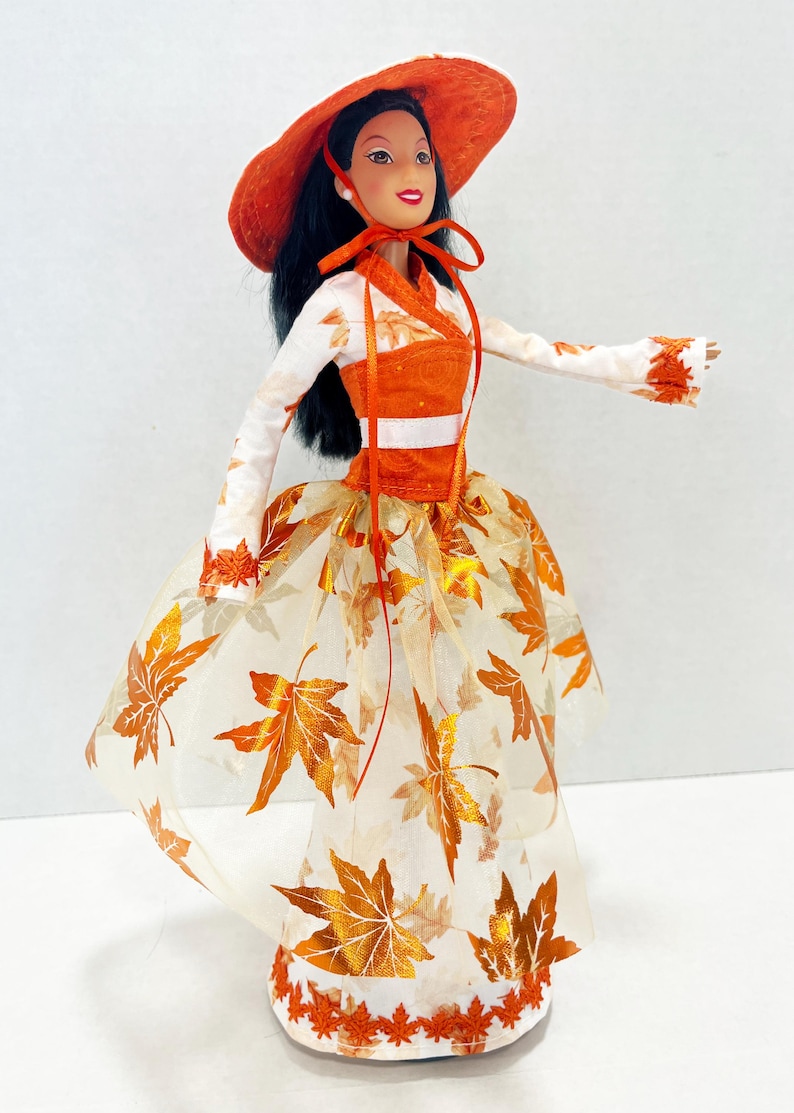 May include: A doll with long black hair wearing an orange wide-brimmed hat and a white long-sleeved top with orange leaf patterns. The outfit includes an orange bodice with a white band and a sheer skirt featuring metallic orange leaf motifs.