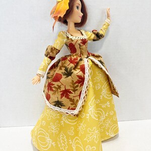 May include: A doll with auburn hair wearing a straw hat adorned with a yellow flower and autumn leaves. The dress features a bodice with a brown and red maple leaf print, a yellow skirt with white pumpkin and leaf outlines, and white lace trim.