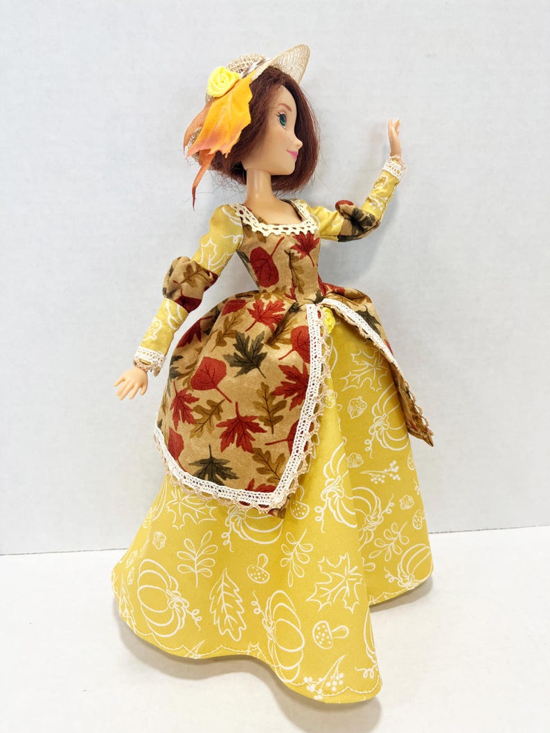 May include: A doll with auburn hair wearing a straw hat adorned with a yellow flower and autumn leaves. The dress features a bodice with a brown and red maple leaf print, a yellow skirt with white pumpkin and leaf outlines, and white lace trim.