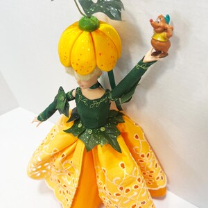 May include: A doll wearing a dark green bodice with leaf accents and a yellow eyelet skirt. The doll has a pumpkin-shaped headpiece with green leaves and holds a small brown mouse figurine with a yellow shirt and blue hat in one raised hand.