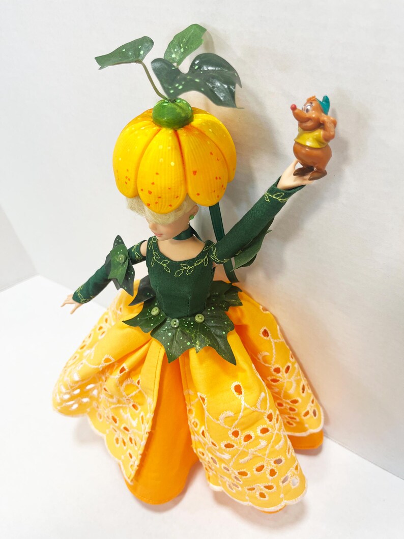 May include: A doll wearing a dark green bodice with leaf accents and a yellow eyelet skirt. The doll has a pumpkin-shaped headpiece with green leaves and holds a small brown mouse figurine with a yellow shirt and blue hat in one raised hand.