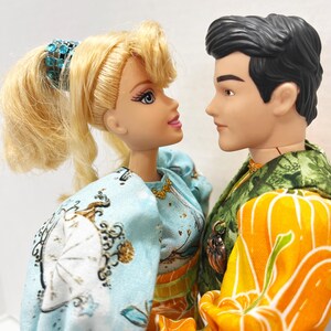 以下が含まれることがあります： 2体の人形がお互いを見つめ合っています。女性の人形はブロンドの髪で、白と金の模様が入った水色のドレスを着ています。男性の人形は、カボチャ模様の緑とオレンジ色のジャケットを着ています。