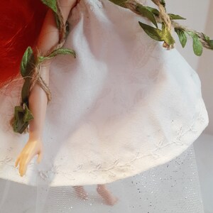 Puede incluir: Un vestido blanco de mu&ntilde;eca con un borde de encaje y una guirnalda de vid verde. El vestido se lleva sobre una falda de tul blanca con purpurina.