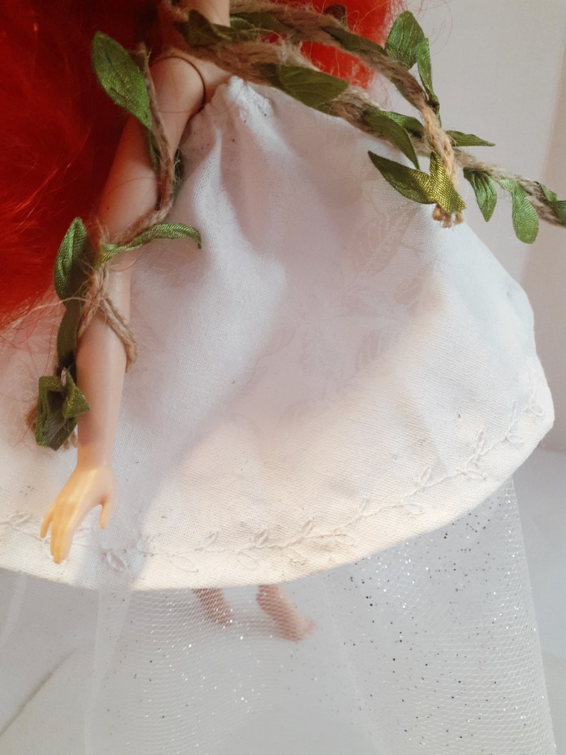 Puede incluir: Un vestido blanco de mu&ntilde;eca con un borde de encaje y una guirnalda de vid verde. El vestido se lleva sobre una falda de tul blanca con purpurina.