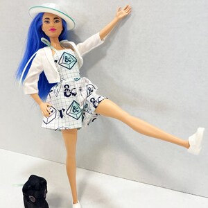 May include: A doll with bright blue hair wearing a white hat, a white cropped jacket, and a white dress featuring a grid pattern with dark blue and light blue geometric shapes. The doll wears white shoes and stands next to a small black animal figure.