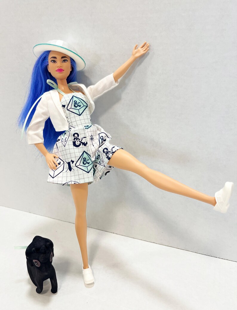 May include: A doll with bright blue hair wearing a white hat, a white cropped jacket, and a white dress featuring a grid pattern with dark blue and light blue geometric shapes. The doll wears white shoes and stands next to a small black animal figure.