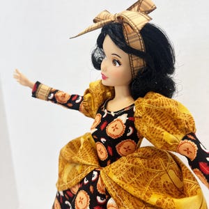 Op de afbeelding: Een pop met zwart haar gestyled met een beige geruite strik-haarband. De pop draagt een jurk van zwarte stof met een herhalend patroon van kleine taartjes en rode appelschijfjes, geaccentueerd door gouden pofmouwen en een gouden taille-overlay.