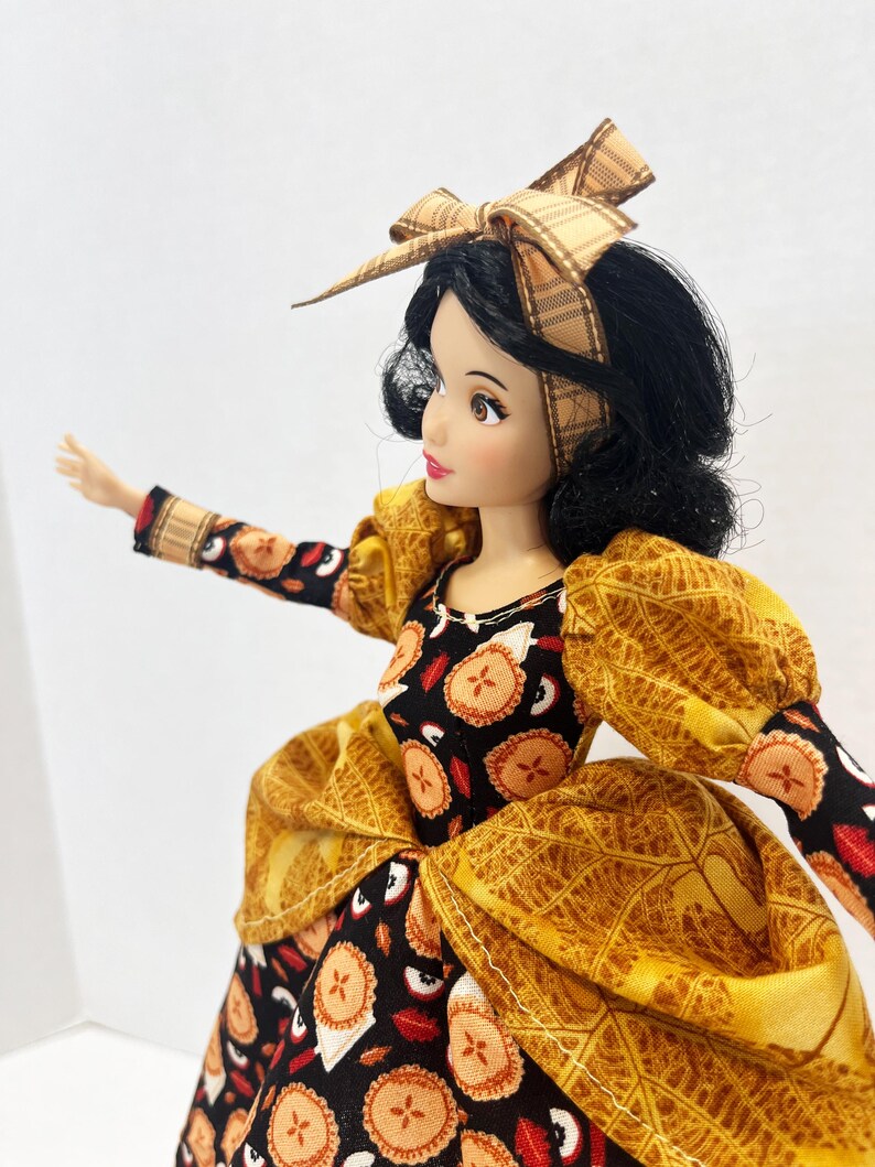 Op de afbeelding: Een pop met zwart haar gestyled met een beige geruite strik-haarband. De pop draagt een jurk van zwarte stof met een herhalend patroon van kleine taartjes en rode appelschijfjes, geaccentueerd door gouden pofmouwen en een gouden taille-overlay.