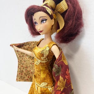 May include: A doll with voluminous dark red hair styled in a high ponytail with a gold ribbon. The doll wears a sleeveless yellow dress featuring a gold leaf pattern and gold leaf appliqu&eacute;s, paired with a cape displaying a fall leaf print.