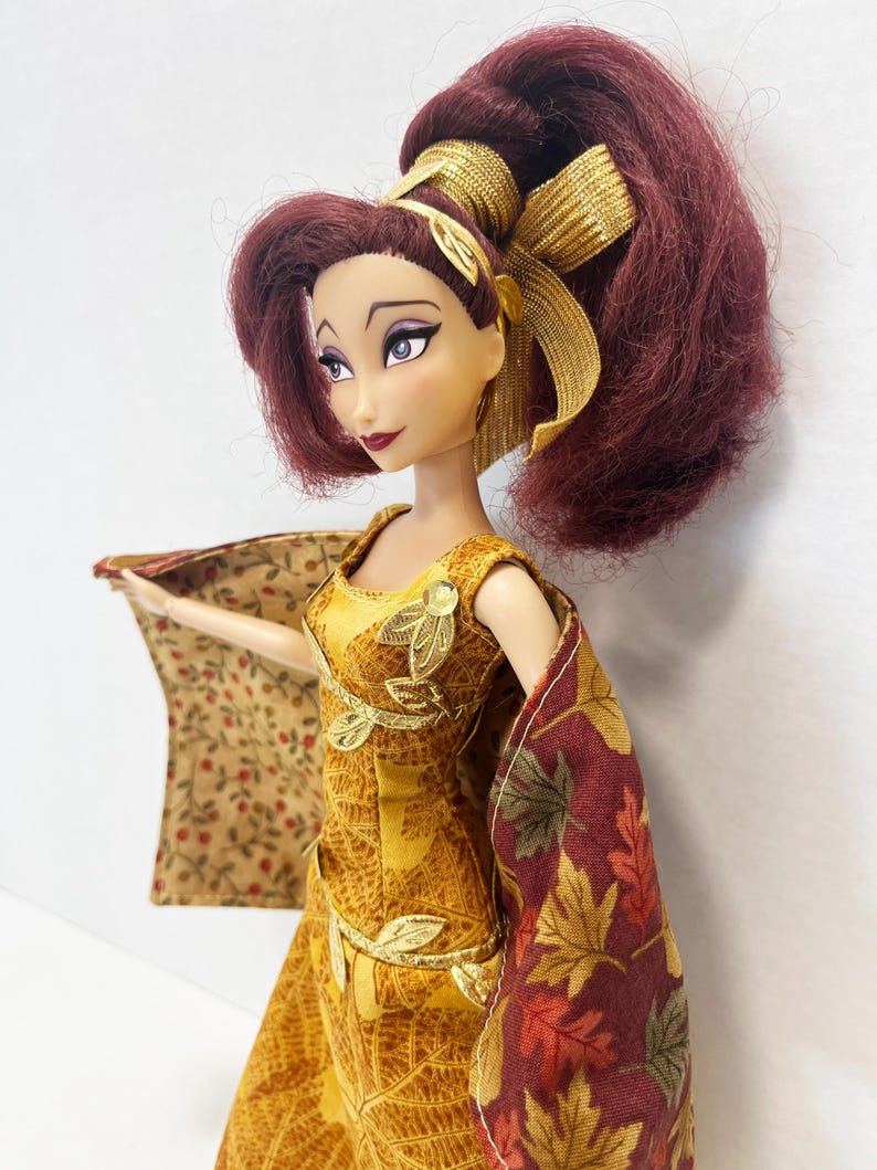 May include: A doll with voluminous dark red hair styled in a high ponytail with a gold ribbon. The doll wears a sleeveless yellow dress featuring a gold leaf pattern and gold leaf appliqu&eacute;s, paired with a cape displaying a fall leaf print.