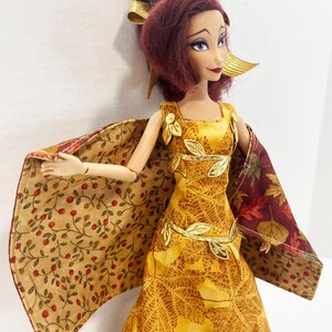May include: A doll with dark red hair styled in an updo with gold ribbons, wearing a yellow dress featuring a leaf pattern and gold leaf accents. The doll has a cape with a tan fabric printed with red berries on one side and a dark autumn leaf print.