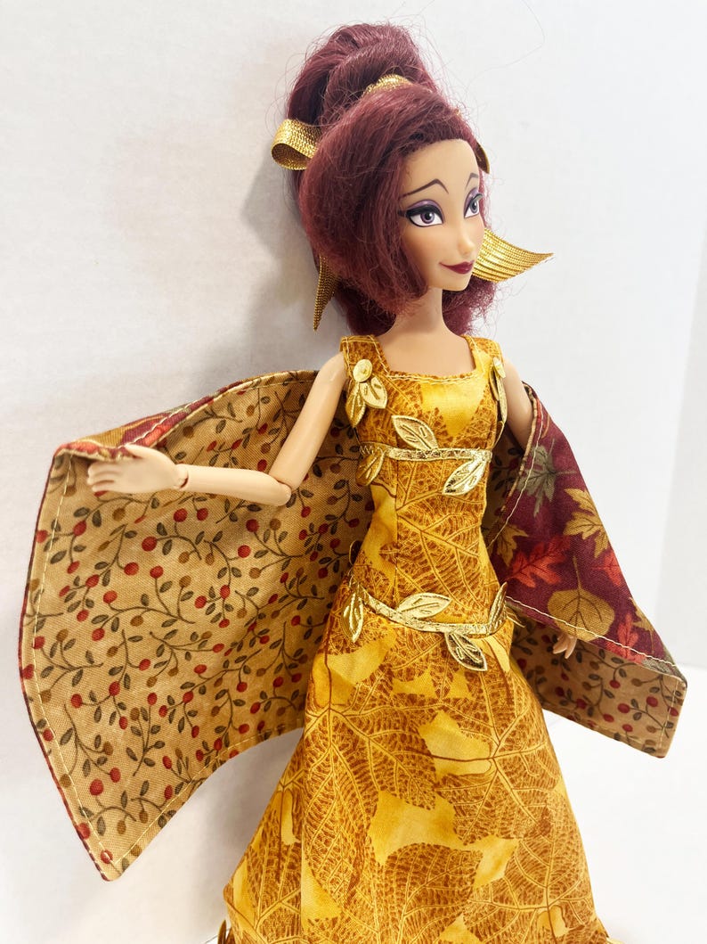 May include: A doll with dark red hair styled in an updo with gold ribbons, wearing a yellow dress featuring a leaf pattern and gold leaf accents. The doll has a cape with a tan fabric printed with red berries on one side and a dark autumn leaf print.