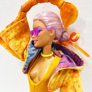 K&ouml;nnte beinhalten: Eine Puppe mit hellrosa Haar, die eine durchscheinende violette Sonnenbrille, ein gelbes Halsband und eine gelbe Kapuzenjacke mit Batikmuster tr&auml;gt. Die Puppe hat ein gro&szlig;es, orangefarbenes, gebogenes Skorpion-Schwanz-Accessoire &uuml;ber dem Kopf.
