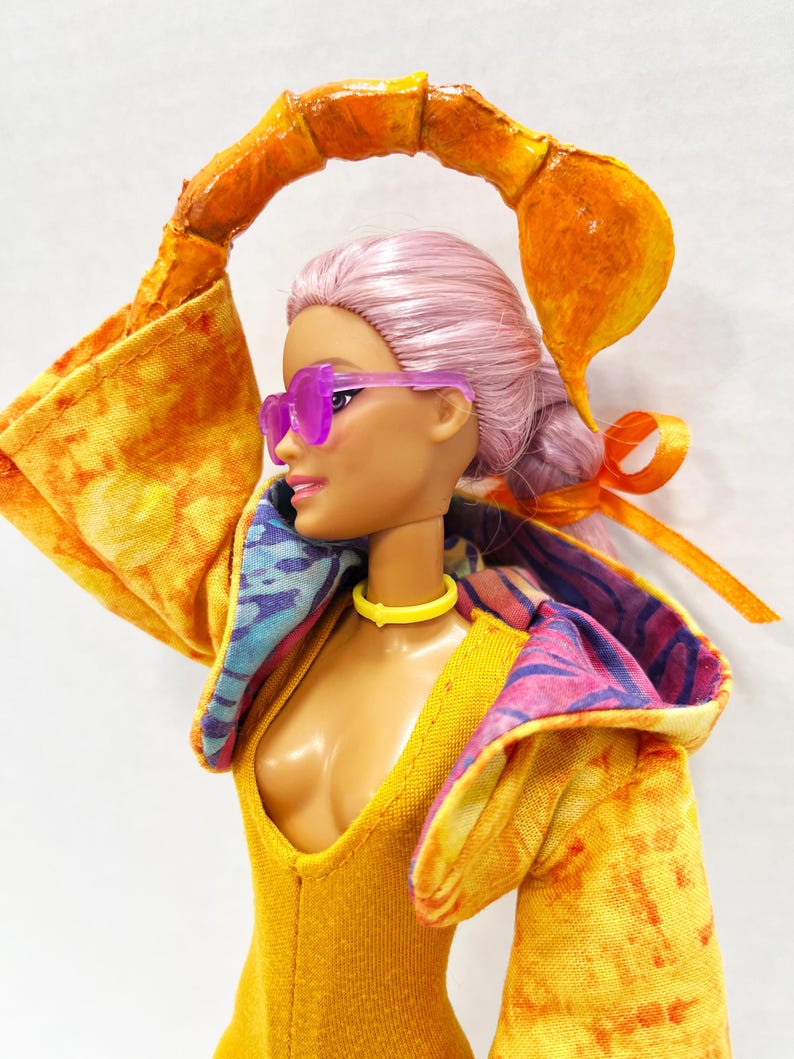K&ouml;nnte beinhalten: Eine Puppe mit hellrosa Haar, die eine durchscheinende violette Sonnenbrille, ein gelbes Halsband und eine gelbe Kapuzenjacke mit Batikmuster tr&auml;gt. Die Puppe hat ein gro&szlig;es, orangefarbenes, gebogenes Skorpion-Schwanz-Accessoire &uuml;ber dem Kopf.
