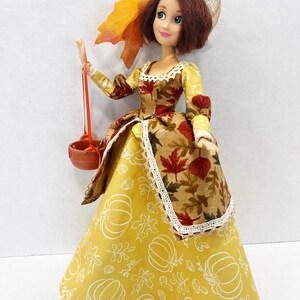 May include: A doll with short brown hair wearing a yellow dress featuring white pumpkin and leaf outlines, paired with a brown overskirt patterned with red and orange autumn leaves. The doll wears a tan mesh hat and carries a small brown basket.