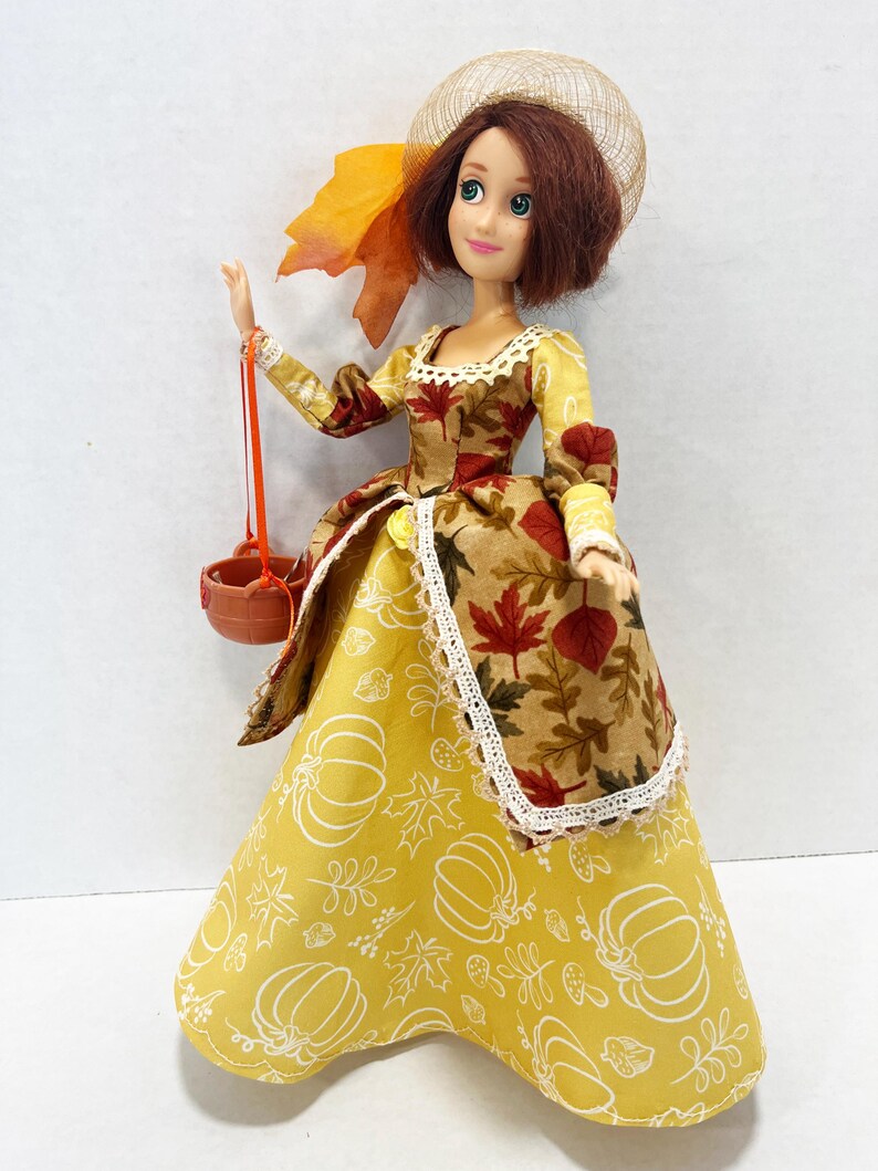 May include: A doll with short brown hair wearing a yellow dress featuring white pumpkin and leaf outlines, paired with a brown overskirt patterned with red and orange autumn leaves. The doll wears a tan mesh hat and carries a small brown basket.