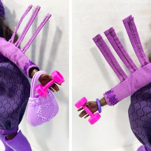 May include: A doll in a purple ensemble, holding a pink dumbbell and a lavender handbag. The doll's outfit includes a long-sleeved top, a skirt with a geometric pattern, and a matching hat. The handbag features a heart-shaped design.