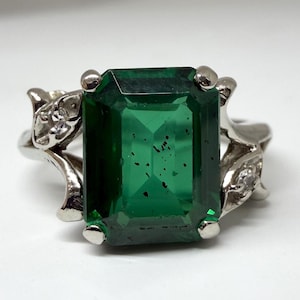 May include: A silver ring featuring a large, emerald-cut green gemstone. The ring has a decorative floral design on the sides with small accent stones. The gemstone is a vibrant green color.