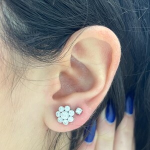 Op de afbeelding: Een paar zilveren oorbelletjes met een cluster van kleine diamanten in de vorm van een bloem. De oorbelletjes worden in de oorlel gedragen.