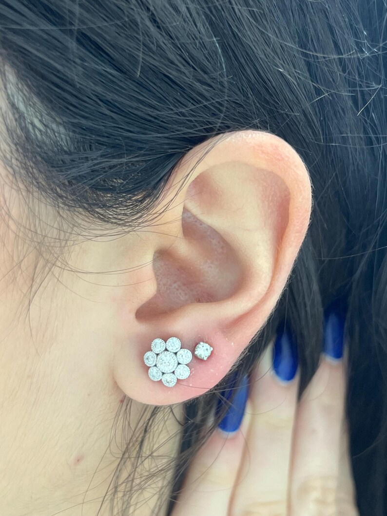 Op de afbeelding: Een paar zilveren oorbelletjes met een cluster van kleine diamanten in de vorm van een bloem. De oorbelletjes worden in de oorlel gedragen.