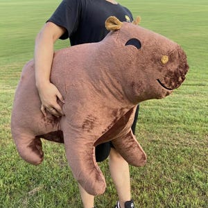 Pode incluir: Um grande bicho de pelúcia de capivara em tons de marrom é segurado por uma pessoa. A capivara tem um rosto sorridente, orelhas pequenas e uma aparência macia e fofa. O fundo é um campo gramado verde.