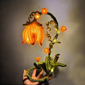 Op de afbeelding: Een grillige, verlichte bloemensculptuur. De grote, oranje bloem is het middelpunt, met een textuur, bloembladachtig ontwerp. De stengel en bladeren zijn groen, met kleine oranje bloemen en kleine lichtjes. De sculptuur wordt door een hand vastgehouden.