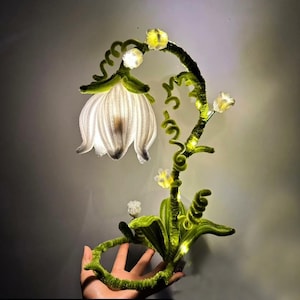 Op de afbeelding: Een decoratieve bloemenlamp met een grote witte bloem en kleinere gele bloemen. De lamp heeft een groene, getextureerde stengel en bladeren, met kleine lampjes die de bloemen verlichten. De lamp wordt door een hand vastgehouden.