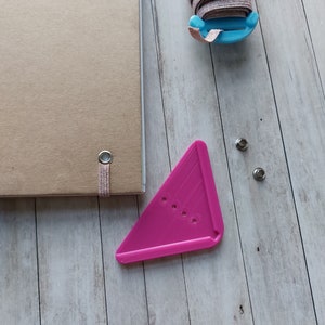 Könnte beinhalten: Ein rosa Plastik-Dreieckswerkzeug mit Löchern zum Stanzen, eine Spule mit rosa Gummiband und zwei silberne Ösen auf einer hellbraunen Holzoberfläche.