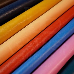 May include: A collection of rolled leather sheets in a variety of colours including dark brown, navy blue, yellow, peach, red, bright blue, light pink, and teal. Each roll displays a distinct textured grain surface arranged in a diagonal pattern.