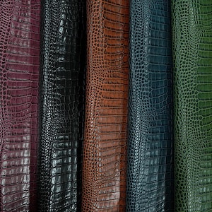 Peut inclure: Sept bandes verticales de cuir présentant un motif texturé de peau de crocodile dans diverses couleurs, dont marron foncé, violet profond, noir, beige, bleu foncé, vert forêt et marron clair, avec un petit logo blanc dans le coin inférieur droit.