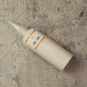 Puede incluir: Una botella blanca con una punta transparente y puntiaguda. La botella está etiquetada con "AL Leather Supply Base Coat".