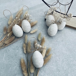 Könnte beinhalten: Fünf weiße Oster-Eier aus Seil, verziert mit getrockneten weißen Blumen und mit Schnur gebunden. Die Eier sind auf einem grauen Hintergrund mit getrocknetem Gras angeordnet.