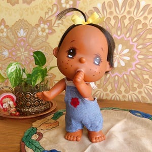 May include: A vinyl doll with dark skin, brown hair tied with a yellow ribbon, and freckles on the cheeks. The doll has large eyes and is wearing blue denim overalls with a small red flower embroidered on the chest, with one hand near the mouth.