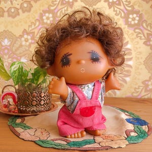 May include: A doll with curly brown hair and a tan complexion, wearing a floral print shirt under pink denim overalls. The doll has painted eyes with long lashes, a small nose, and an open mouth. Its arms are raised, and it has bare feet.