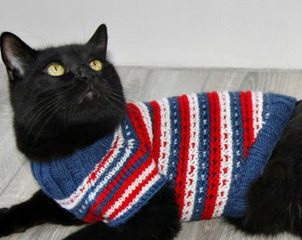 May include: A black cat wearing a blue, white, and red striped knitted sweater. The cat is lying on a light gray wood floor.