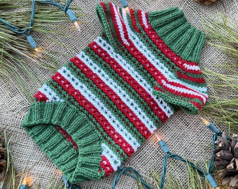 May include: A green, red, and white striped knitted sweater for a pet. The sweater is laid out on a burlap background with pine needles and Christmas lights.