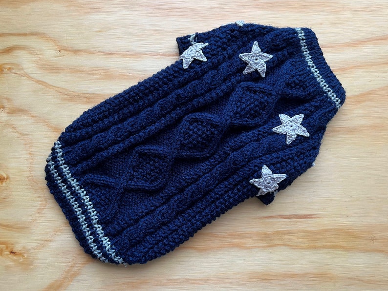 K&ouml;nnte beinhalten: Ein dunkelblauer gestrickter Hundepullover mit Zopfmuster und wei&szlig;en Stern-Applikationen.