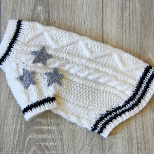 K&ouml;nnte beinhalten: Wei&szlig;er gestrickter Hundepullover mit schwarzen Streifen und grauen Stern-Applikationen.