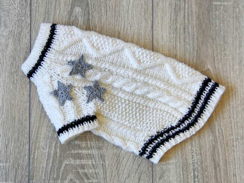 K&ouml;nnte beinhalten: Wei&szlig;er gestrickter Hundepullover mit schwarzen Streifen und grauen Stern-Applikationen.