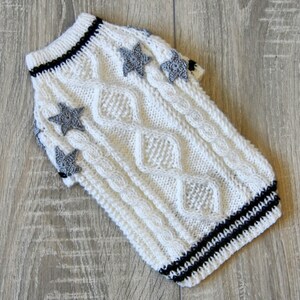 May include: White knitted dog sweater with a cable knit pattern and gray star embellishments. The sweater has black and white stripes around the neck and sleeves.