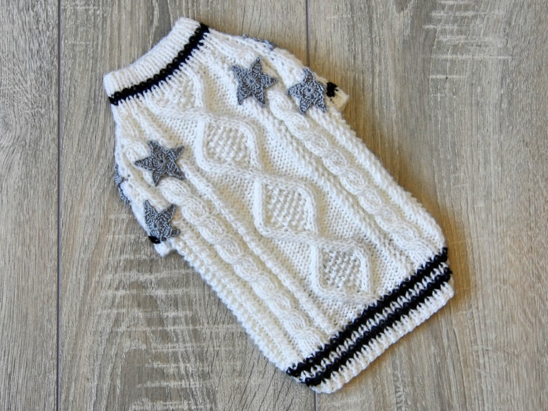 May include: White knitted dog sweater with a cable knit pattern and gray star embellishments. The sweater has black and white stripes around the neck and sleeves.